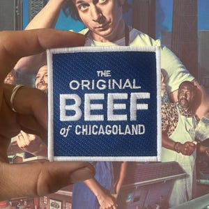 Puede incluir: Parche cuadrado con el texto "THE ORIGINAL BEEF of CHICAGOLAND" bordado en blanco sobre un fondo azul. El parche se sostiene frente a una imagen de fondo de personas.
