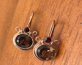 May include: A pair of rose gold earrings. Each earring features a large, oval-cut smoky quartz gemstone, a small, round garnet, and decorative swirls. The earrings are displayed on a wooden surface.