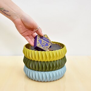 May include: Three stacked geometric bowls in blue, green, and yellow. The bowls are made of a textured material and have a unique, modern design. A small purple box with gold lettering that reads "Chocolate Frog" is sitting in the top bowl.
