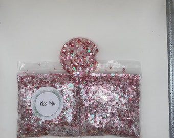 Op de afbeelding: Een ronde auto-onderzetter van hars en twee doorzichtige plastic zakjes gevuld met roze, rode en witte hartvormige glitters. Het linkerzakje heeft een rond label met de tekst Kiss Me en een klein logo met de tekst The Wright Glitter.