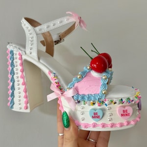 May include: White platform heels with a pink and blue frosting design, topped with cherries and sprinkles. The heels have a white strap with pearl accents and a pink bow. The platform features two candy hearts that say "Be Mine".