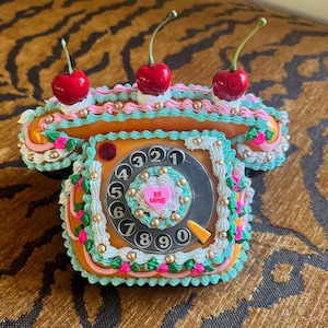 May include: A whimsical, orange-colored telephone-shaped cake decorated with pink, teal, and white frosting. The cake is topped with three cherries and gold sprinkles. The dial features numbers and a heart-shaped candy that says "BE MINE".