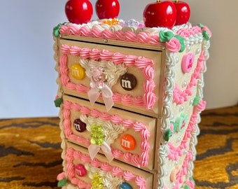 May include: A whimsical, decorative jewelry box resembling a cake, adorned with pink frosting, white icing, and red cherries. The box features three drawers decorated with M&M's candies and bows, creating a playful, unique design.