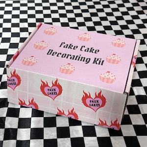 Può includere: Una scatola rosa con la scritta "Fake Cake Decorating Kit" e illustrazioni di cupcake. La scatola presenta un motivo ripetuto di cuori fiammeggianti con la scritta "Faux Cakes". Lo sfondo è un motivo a scacchiera in bianco e nero.