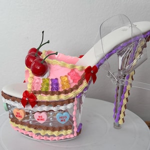 May include: A novelty high heel shoe designed to look like a cake. The shoe has a white sole, clear straps, and a clear platform base decorated with pink, yellow, and brown frosting. It is decorated with gummy bears, cherries, and heart-shaped candies with the words "Be Mine" and "Hug Me".