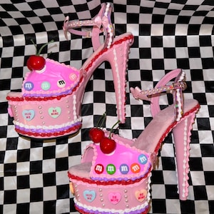 May include: Pink platform high heels with a cake design. The heels feature a cherry on top, M&M candies, and heart-shaped candies with phrases like "HUG ME" and "BE MINE." The heels have a strap and a high stiletto heel.