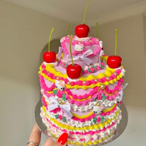 Peut inclure: Un gâteau à deux étages rose et jaune décoré de cerises, de nœuds blancs et de personnages Hello Kitty. Le gâteau est sur un support à gâteau argenté.