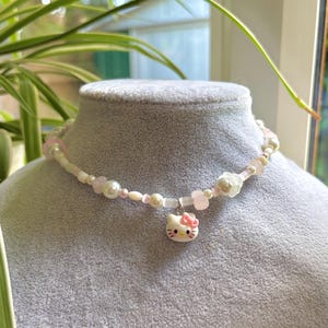 May include: A beaded necklace featuring a variety of white and light pink beads, including round pearls and translucent shapes. A small, white Hello Kitty charm with a pink bow hangs from the centre of the strand, secured by a silver-toned metal loop.