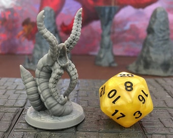 Puede incluir: Una miniatura gris y detallada de una criatura con forma de gusano con tentáculos y una boca abierta. La miniatura está sobre una base gris. Un dado amarillo de 20 caras con números negros está en el fondo.