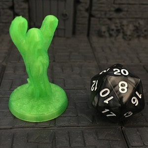 Może przedstawiać: Zielona plastikowa miniatura stworzenia z uniesionymi ramionami, stojąca na okrągłej podstawie. Czarna 20-ścienna kostka do gry z białymi liczbami jest w tle.