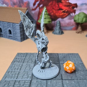 May include: A gray skeletal dragon miniature with wings outstretched, standing on a gray and white tiled surface. A bright orange 20-sided die is in the foreground.