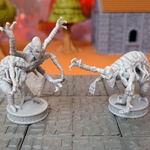 May include: Two gray, detailed, multi-legged, humanoid figures with long arms and claws. The figures are standing on round bases with the names "Jim Lovell" and "Wind Like" printed on them.