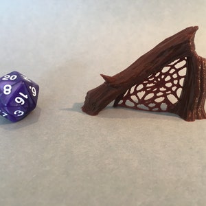 May include: A brown, 3D printed miniature of a spider web stretched between two tree branches. The web is detailed and intricate. A purple 20-sided die is in the foreground.