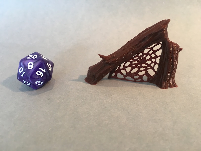May include: A brown, 3D printed miniature of a spider web stretched between two tree branches. The web is detailed and intricate. A purple 20-sided die is in the foreground.