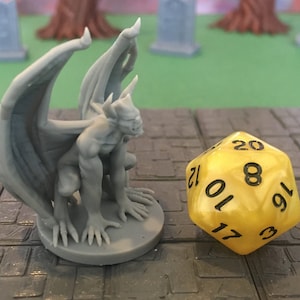 Puede incluir: Una g&aacute;rgola miniatura de pl&aacute;stico gris con alas, sentada sobre una base gris. La g&aacute;rgola est&aacute; mirando hacia la izquierda y tiene una escultura detallada. Un dado de 20 caras amarillo con n&uacute;meros negros est&aacute; en primer plano.