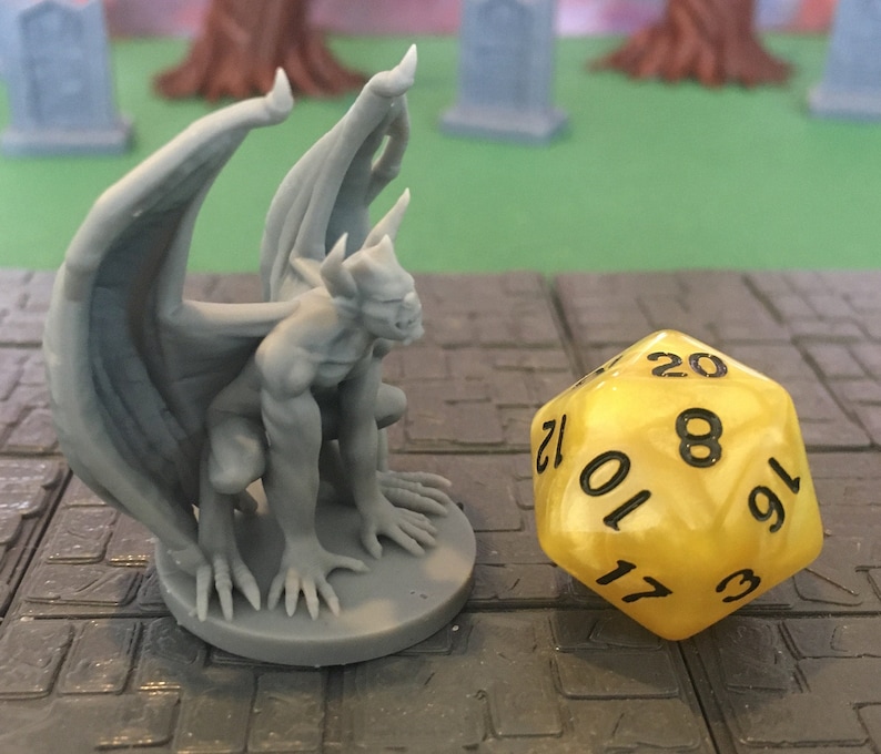 Puede incluir: Una g&aacute;rgola miniatura de pl&aacute;stico gris con alas, sentada sobre una base gris. La g&aacute;rgola est&aacute; mirando hacia la izquierda y tiene una escultura detallada. Un dado de 20 caras amarillo con n&uacute;meros negros est&aacute; en primer plano.