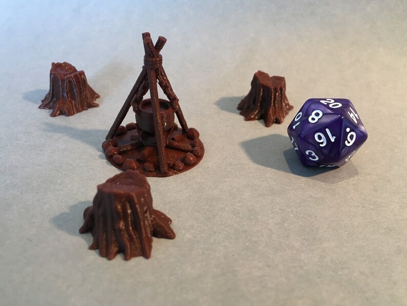 May include: A purple 20-sided die with white numbers sits on a table next to a brown 3D printed miniature of a campfire with a cauldron and three tree stumps.