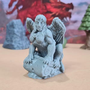May include: A gray figurine of a sphinx with wings, seated on a stone base. The sphinx has a human face and a lion's body. The figurine is likely for tabletop gaming or display.