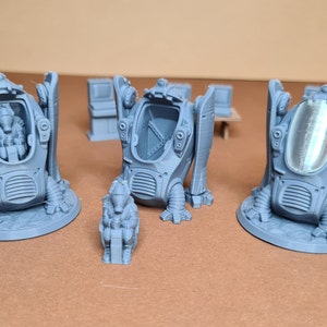 May include: Three grey 3D printed miniature figures of a futuristic vehicle with a transparent dome. The vehicle has a detailed design with a textured surface and a base. The figures are on a brown surface.