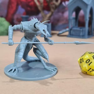 May include: A gray, 3D-printed miniature of a reptilian humanoid creature holding a spear, posed in a combat stance. A yellow, multi-sided die with black numbers sits nearby. The miniature is on a round base.