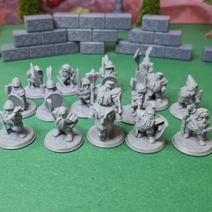 May include: A collection of grey miniature fantasy figures, including dwarves, on a green surface. The figures are in various poses, some holding weapons and shields. Grey stone block structures are in the background.