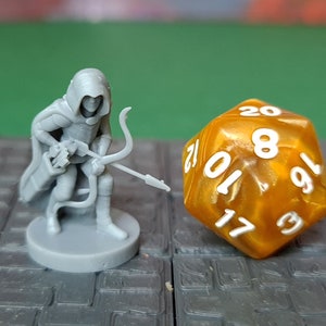 Puede incluir: Una figura en miniatura de plástico gris de una figura encapuchada que sostiene un arco y una flecha. La figura está de pie sobre una base gris. Un dado de 20 caras dorado con números blancos está en el fondo.