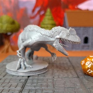 Può includere: Una statuetta di dinosauro grigio con bocca aperta, denti affilati e texture della pelle dettagliata. La statuetta è su una base rotonda con testo e accanto a un dado dorato a più facce. La scena è ambientata su una superficie piastrellata grigia.