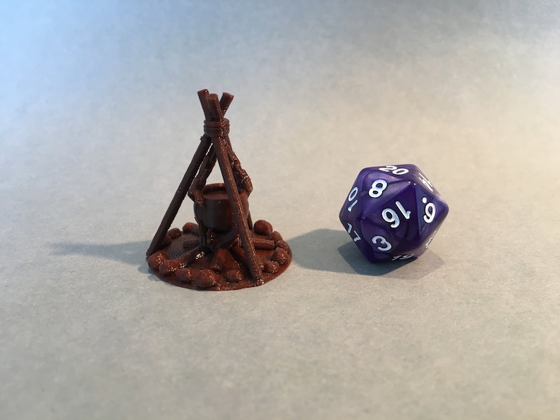 May include: A brown 3D printed miniature of a campfire with a cauldron hanging over the flames. A purple 20-sided die with white numbers is in the background.