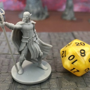 Puede incluir: Una figura en miniatura de plástico gris de un elfo mago sosteniendo un bastón. El elfo lleva una túnica y tiene orejas puntiagudas. La figura está de pie sobre una base gris. Un dado de 20 caras amarillo está en el fondo.