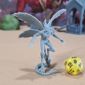Può includere: Una miniatura grigia e dettagliata di una creatura umanoide alata con artigli affilati e una coda serpentina. Un dado giallo a venti facce è accanto alla figura. Altre strutture in miniatura sono sullo sfondo.