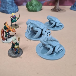 Könnte beinhalten: Miniaturfiguren von Fantasy-Charakteren und -Kreaturen. Die Szene zeigt einen Krieger mit Bogen, eine Figur mit Flammen und mehrere graue frosch- und leguanartige Kreaturen auf kreisförmigen Sockeln. Der Hintergrund zeigt Miniatur-Gelände.