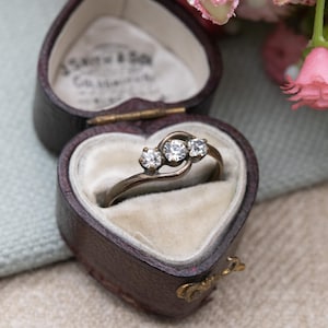 May include: A vintage ring with three clear gemstones set in a swirling design. The ring is displayed in a heart-shaped, brown leather jewelry box with a cream-colored interior. The box is open, revealing the ring.