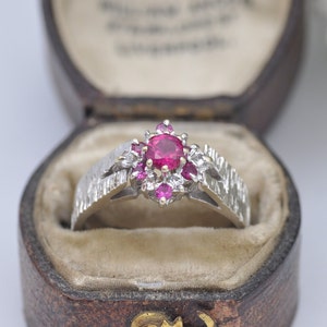 May include: A silver ring with a large red ruby surrounded by smaller pink and white stones. The ring is in a brown velvet ring box.