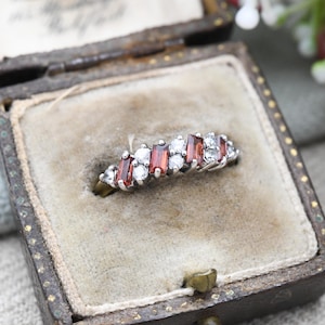 May include: A silver ring with alternating rectangular red and round white gemstones. The ring is displayed inside a vintage, brown jewelry box with a beige interior.
