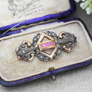 Może przedstawiać: Antyczna broszka z centralną prostokątną złotą oprawą z różowymi kamieniami szlachetnymi, obramowaną romboidalnym wzorem. Broszka posiada bogate zdobienia ze srebra i złota, umieszczona w fioletowym pudełku wyłożonym aksamitem.