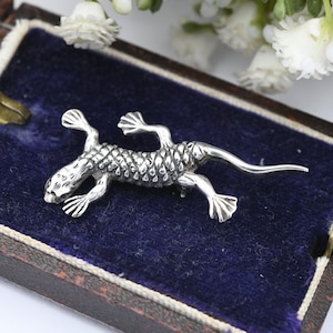 May include: A silver lizard brooch with detailed scales and limbs, presented in a vintage jewellery box with a blue velvet lining. The brooch's design is intricate, highlighting the craftsmanship of the reptile-themed accessory.