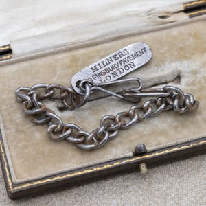 May include: An antique silver-toned chain bracelet with a matching tag. The tag reads "MILNERS FINSBURY PAVEMENT LONDON". The bracelet is displayed in a vintage beige jewelry box with a gold trim.
