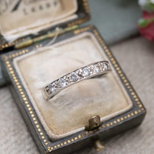 Op de afbeelding: Een zilveren ring met een rij kleine, heldere edelstenen. De ring wordt gepresenteerd in een vintage sieradendoosje met een donkergroene buitenkant, gouden randen en een zachte, fluwelen voering.