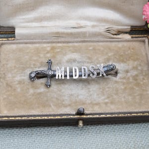 Può includere: Spilla color argento a forma di spada, con la scritta "MIDDSX" in maiuscolo. La spilla è esposta all'interno di un cofanetto vintage foderato in velluto. La scatola è beige e ha un bordo verde scuro.