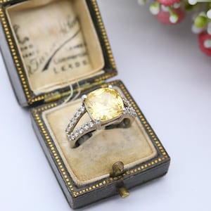May include: A vintage ring with a large, square, yellow gemstone set in a silver band, accented with smaller clear stones. The ring is displayed in a black and gold jewelry box with a beige interior.