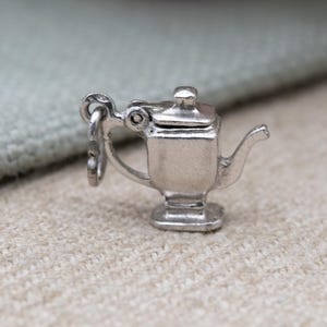 Könnte beinhalten: Ein kleiner, silberfarbener Teekannen-Anhänger mit einer Schlaufe zum Befestigen an einem Armband oder einer Halskette. Die Teekanne hat einen quadratischen Sockel, einen gebogenen Ausguss und einen kleinen Deckel. Der Anhänger ist detailliert.
