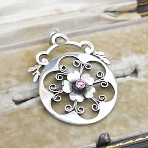 Puede incluir: Colgante de plata con diseño floral. El colgante presenta una flor central con una gema rosa y está rodeado por un diseño calado con detalles en espiral. El colgante se muestra sobre una superficie de terciopelo.