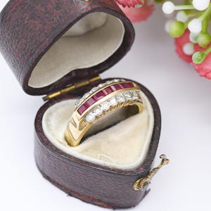 May include: A gold ring featuring a line of rectangular red gems and a row of clear, round gemstones. The ring is presented in a heart-shaped, burgundy leather jewellery box with a cream interior.
