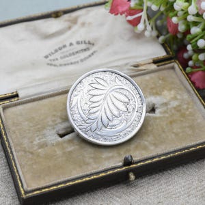 May include: A round, silver-toned brooch with an engraved floral design, displayed in a vintage jewelry box. The brooch features a detailed pattern of leaves and curves. The box is lined with beige velvet and has gold trim.