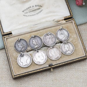 Może przedstawiać: Kolekcja antycznych srebrnych wisiorków przedstawiających profile postaci historycznych, prezentowanych w zabytkowym pudełku. Wisiorki są okrągłe z małą pętelką do zawieszenia. Pudełko jest wyłożone kremową tkaniną i ma złote obramowanie. Widoczny jest napis "WILSON & GILL, THE GOLDSMITHS".