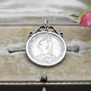 May include: A silver pendant with a detailed profile of a crowned woman, possibly a historical figure. The pendant has decorative scrollwork at the top and is set against a soft, neutral background. The coin-like design suggests a vintage or antique piece.