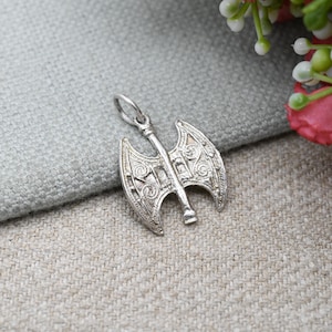 以下が含まれることがあります： 精巧な彫刻が施されたシルバーカラーのアックスペンダント。ペンダントは、チェーン用のループが付いた両刃のアックスデザインが特徴です。アックスは、グレーのテクスチャード加工された表面に表示され、背景にはぼやけた花の配置があります。