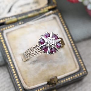 Op de afbeelding: Een zilveren ring met een cluster van rode robijnen en een enkele diamant in het midden. De ring is gezet op een getextureerde band.