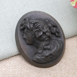 Puede incluir: Cameo ovalado gris oscuro con un perfil femenino clásico con detalles de cabello y acentos de vid. El cameo se encuentra sobre un fondo neutro, resaltando el intrincado tallado en relieve.