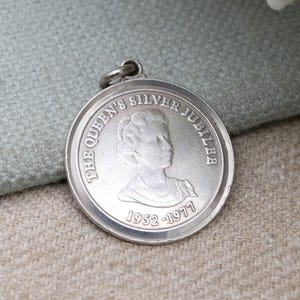 Op de afbeelding: Een zilveren hanger met een cirkelvormig ontwerp. De hanger toont een profiel van een persoon en de tekst "THE QUEEN'S SILVER JUBILEE 1952-1977" rondom de rand. Het heeft een kleine lus aan de bovenkant.